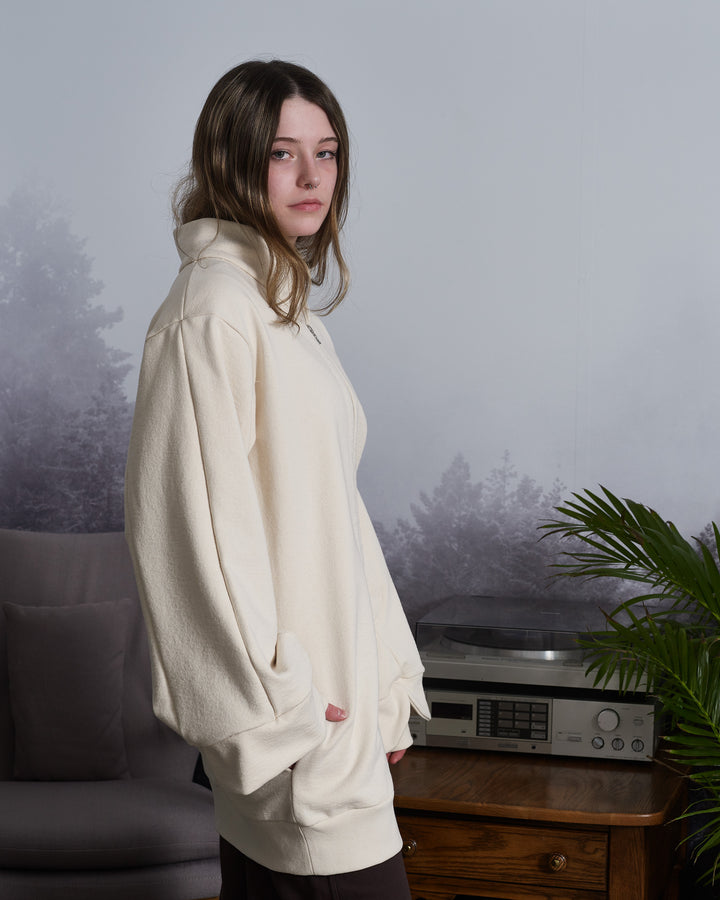 A girl wearing a beige, oversized tulip-shaped sleeve coat with a rib cuff and a matching petal collar, featuring a 2-way riri metal zipper and side pockets.