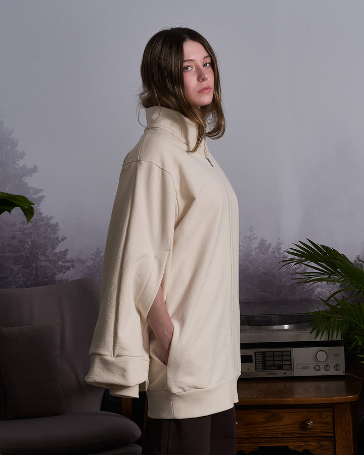 A girl weraing a beige, oversized tulip-shaped sleeve coat with a rib cuff and a matching petal collar, featuring a 2-way riri metal zipper and side pockets.