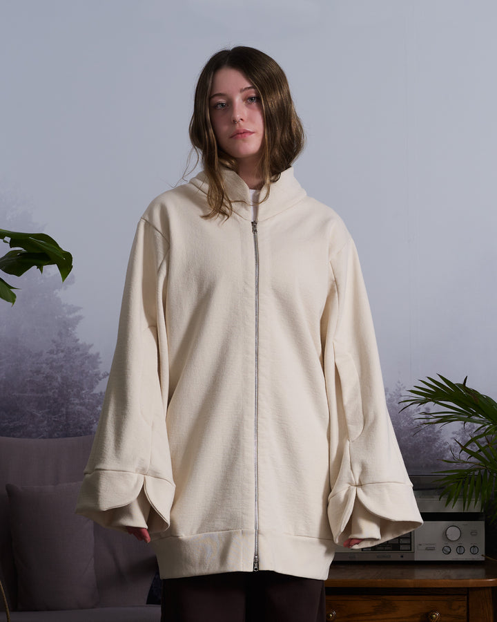 A girl wearing a beige, oversized tulip-shaped sleeve coat with a rib cuff and a matching petal collar, featuring a 2-way riri metal zipper and side pockets.