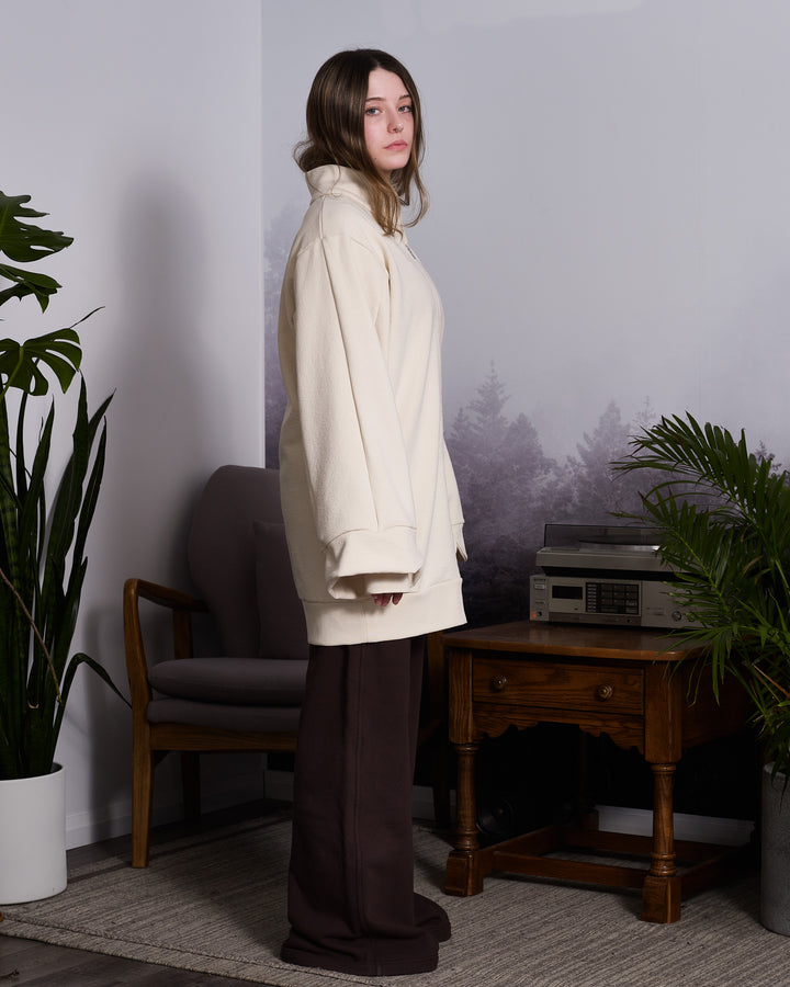 A girl wearing wide leg brown sweat pants and a beige, oversized tulip-shaped sleeve coat with a rib cuff and a matching petal collar, featuring a 2-way riri metal zipper and side pockets.