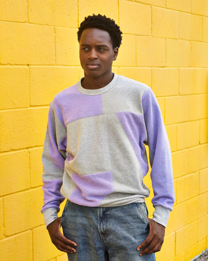 A boy standing infornt of a yellow brick wall. He is wearing jeans and an upcycled crewneck sweater with heather grey and lavender pieces combined to make a unique geometric pattern.