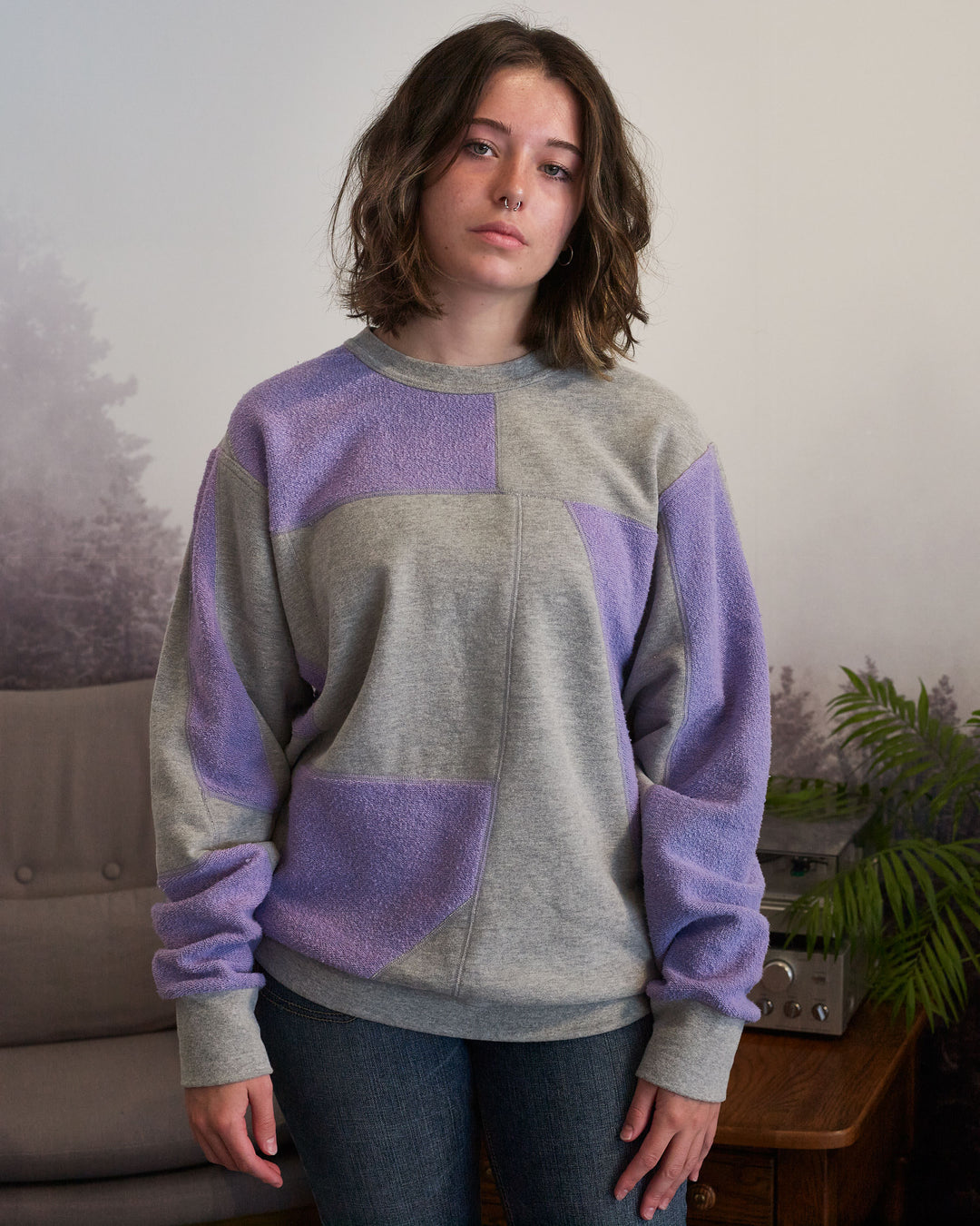 A girl standing inside a living room. She is wearing jeans and an upcycled crewneck sweater with heather grey and lavender pieces combined to make a unique geometric pattern.