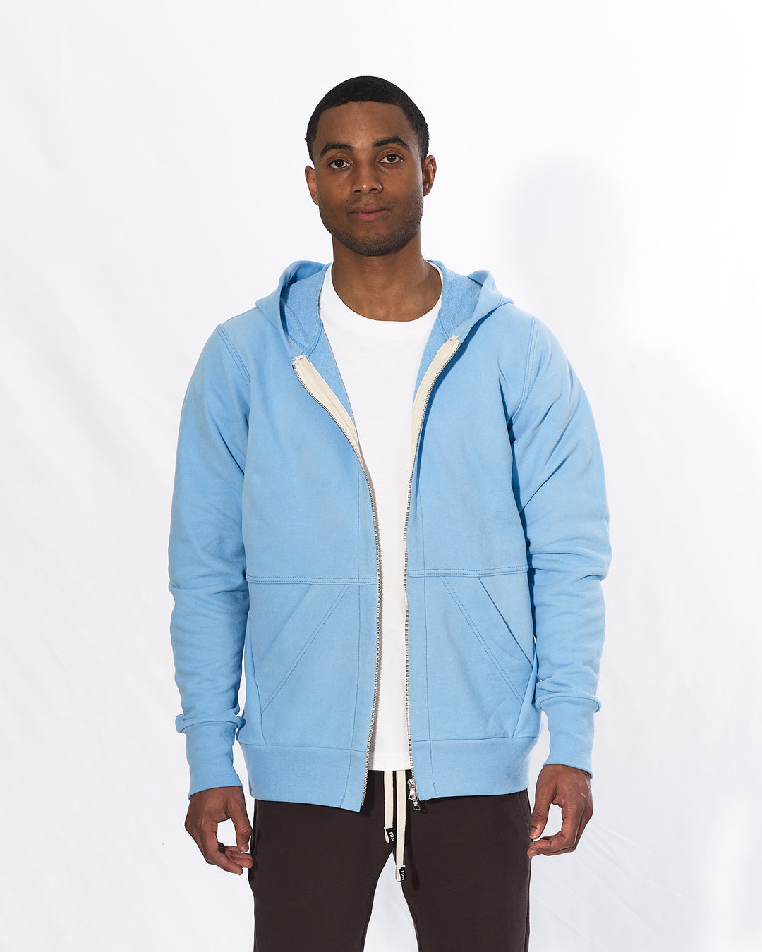 A man wearing a light blue zip-up hoodie with brown pants in front of a white background. The hoodie has a two way silver zipper and two front pockets.