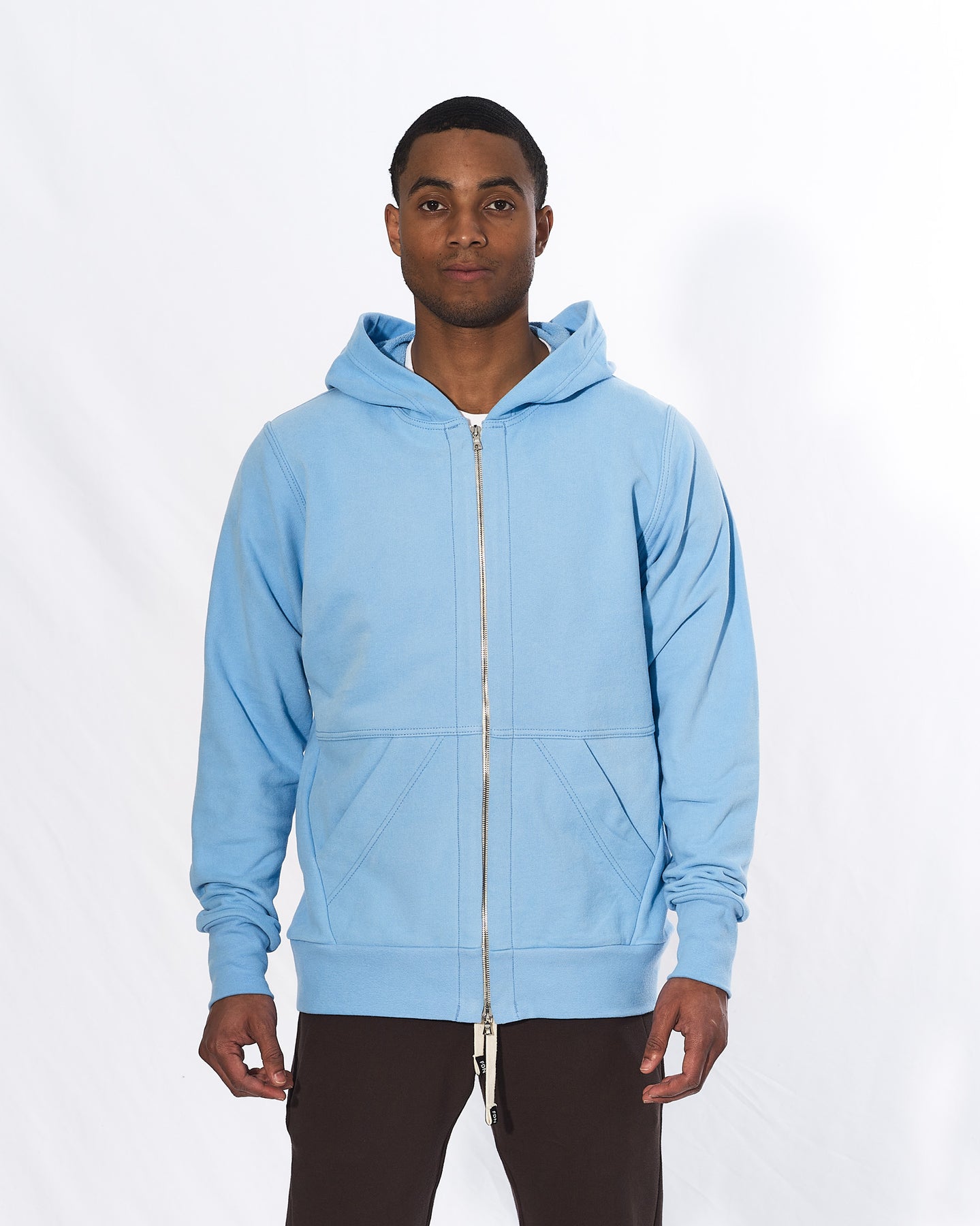 A man wearing a light blue zip-up hoodie with brown pants in front of a white background. The hoodie has a two way silver zipper and two front pockets.