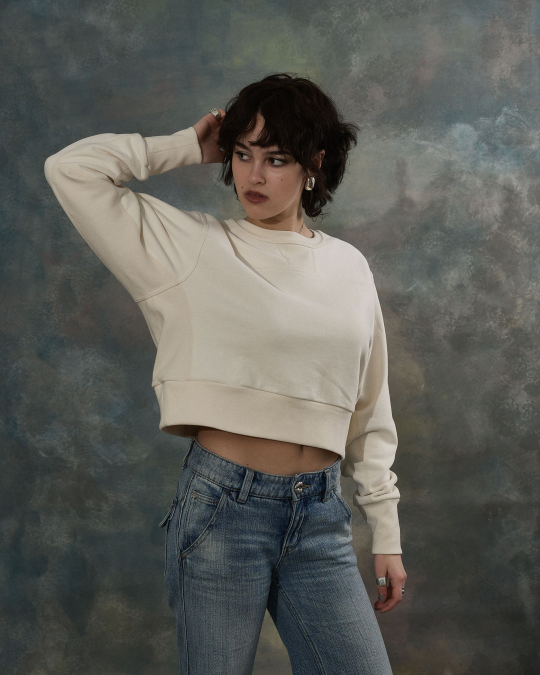 Person wearing a beige cropped sweatshirt and blue jeans against a textured gray background