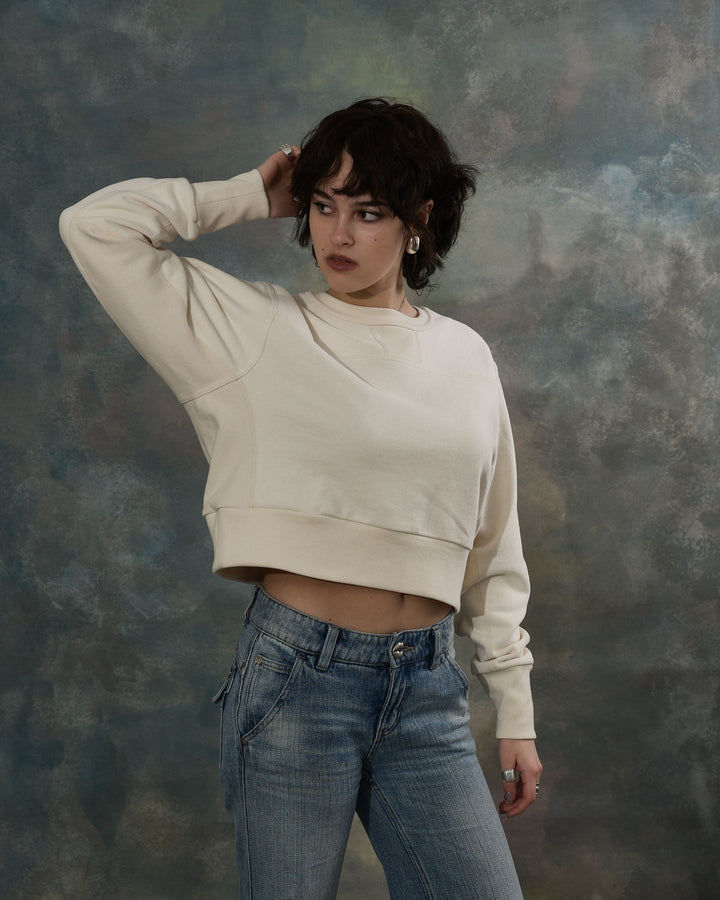Person wearing a beige cropped sweatshirt and blue jeans against a textured gray background