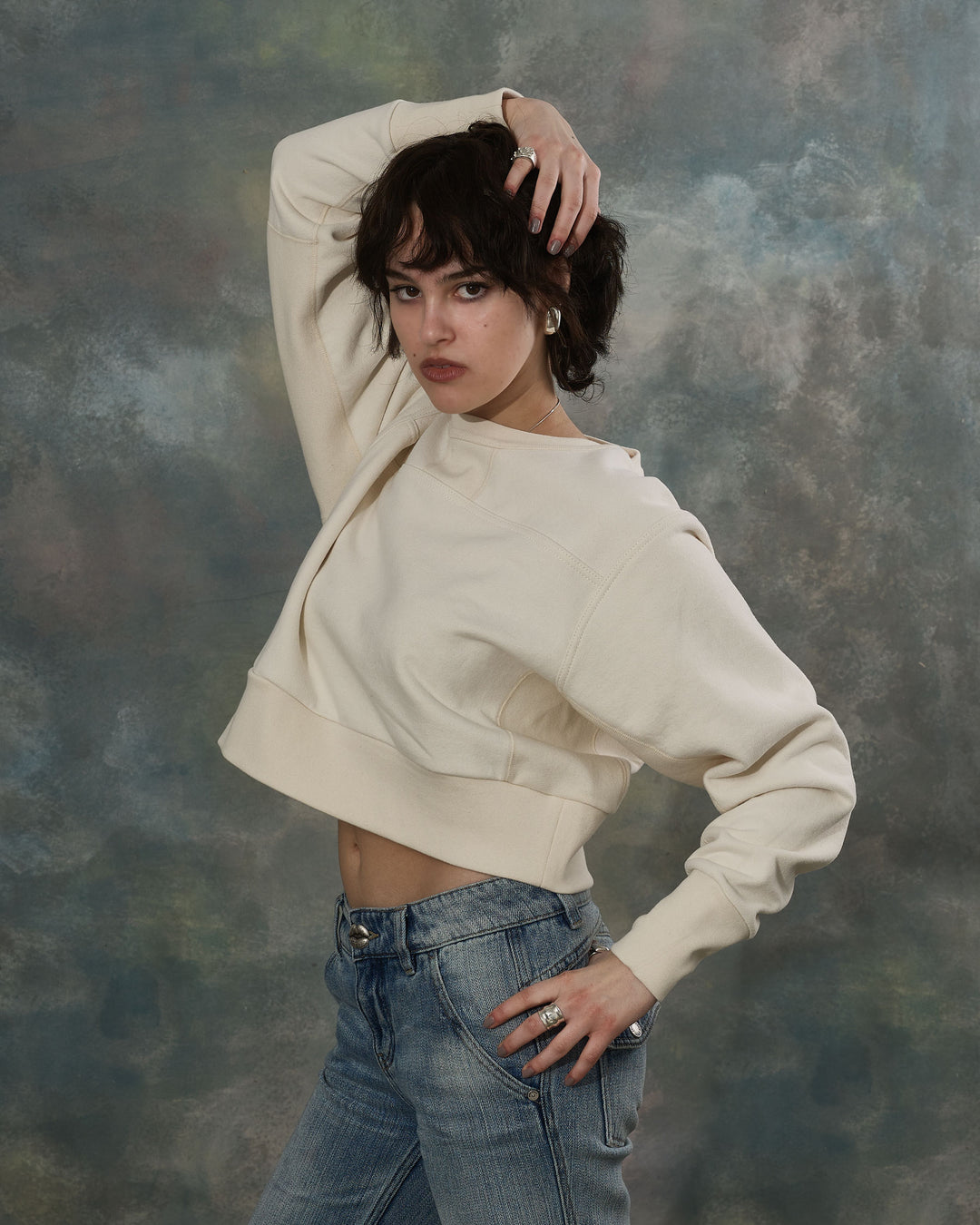 Person wearing a beige cropped sweatshirt and blue jeans against a textured gray background