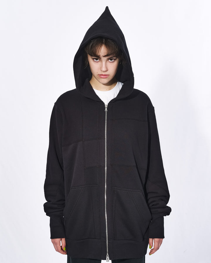 A girl with short black hair standing in front of a white background. She is wearing an all black oversized upcycled hoodie with a silver zipper. Her hood is on her head and pointed at the top.