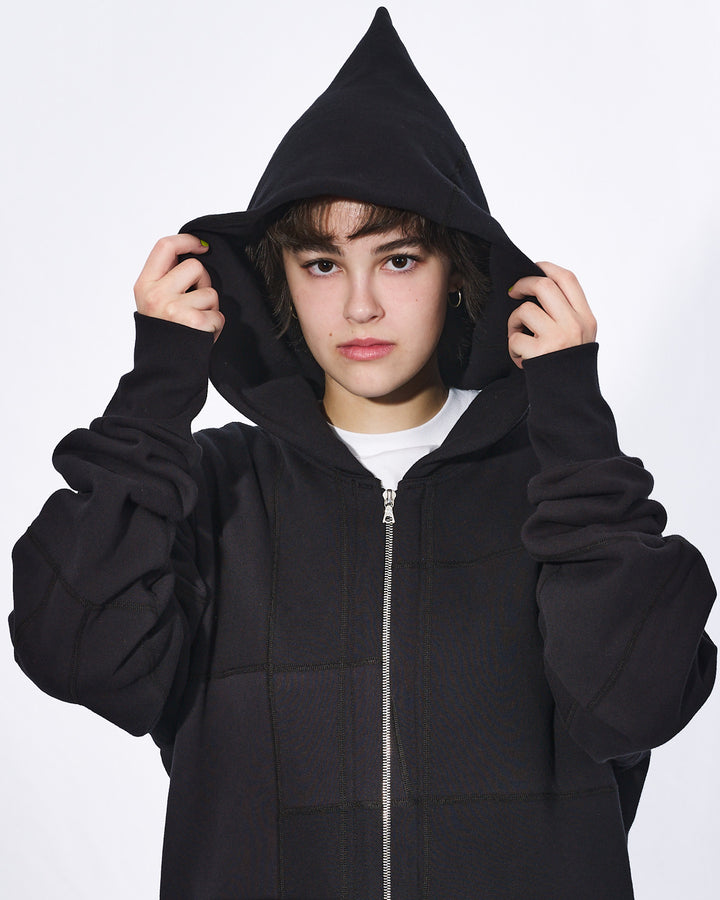 A close up of girl with short black hair with her hood up wearing an all black oversized upcycled hoodie with a silver zipper.