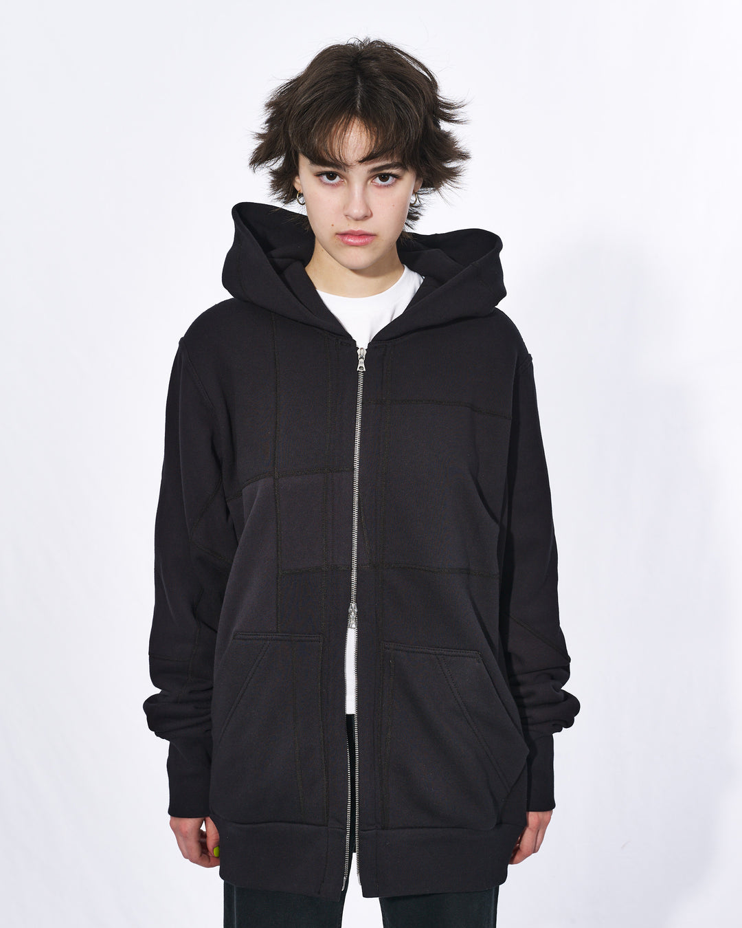 A girl with short black hair wearing an all black oversized upcycled hoodie with a silver two way zipper and two front pockets.