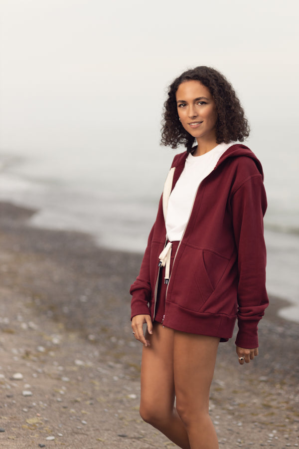 A girl with curly shoulder length brown hair standing at the shoreline on a rock beach in overcast weather. She is wearing a maroon zip-up hoodie, white t-shirt and short maroon shorts. The hoodie has a two way silver zipper and two front pockets.