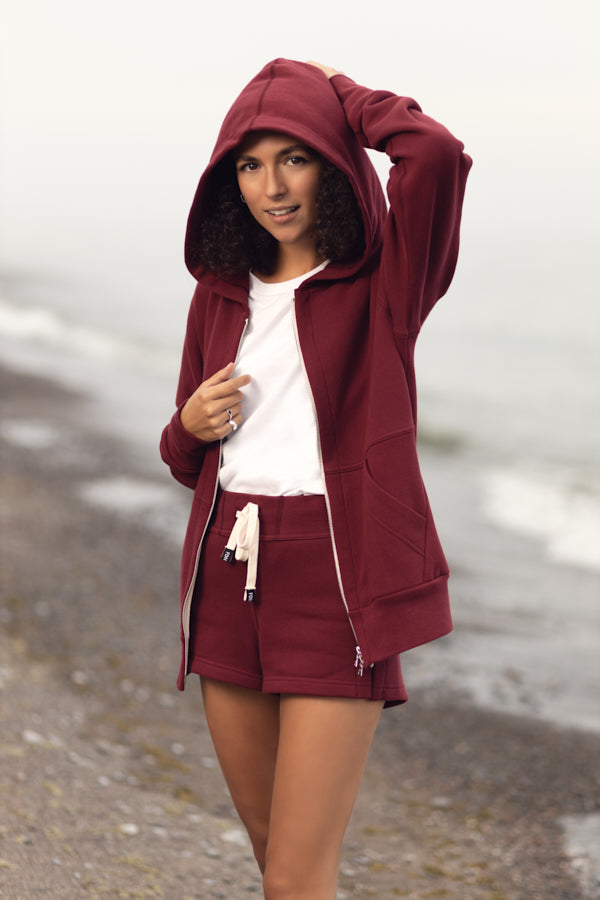 A girl with curly shoulder length brown hair standing at the shoreline on a rock beach in overcast weather. She is wearing a maroon zip-up hoodie with the hood up, a white t-shirt and short maroon shorts with white drawstring. The hoodie has a two way silver zipper and two front pockets.