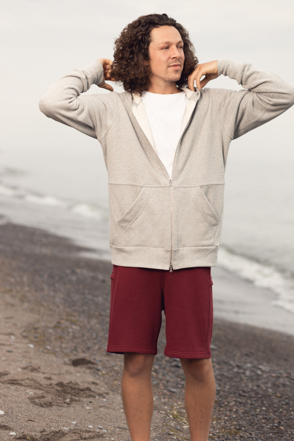 A man with curly long hair standing on the beach. Wearing a grey zip-up hoodie and maroon shorts. The hoodie had a two way silver zipper and two front pockets.