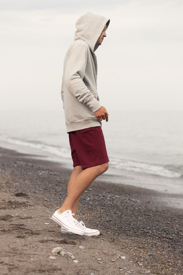 A man walking on the beach in overcast weather. Wearing a grey zip-up hoodie, maroon shorts and white shoes.