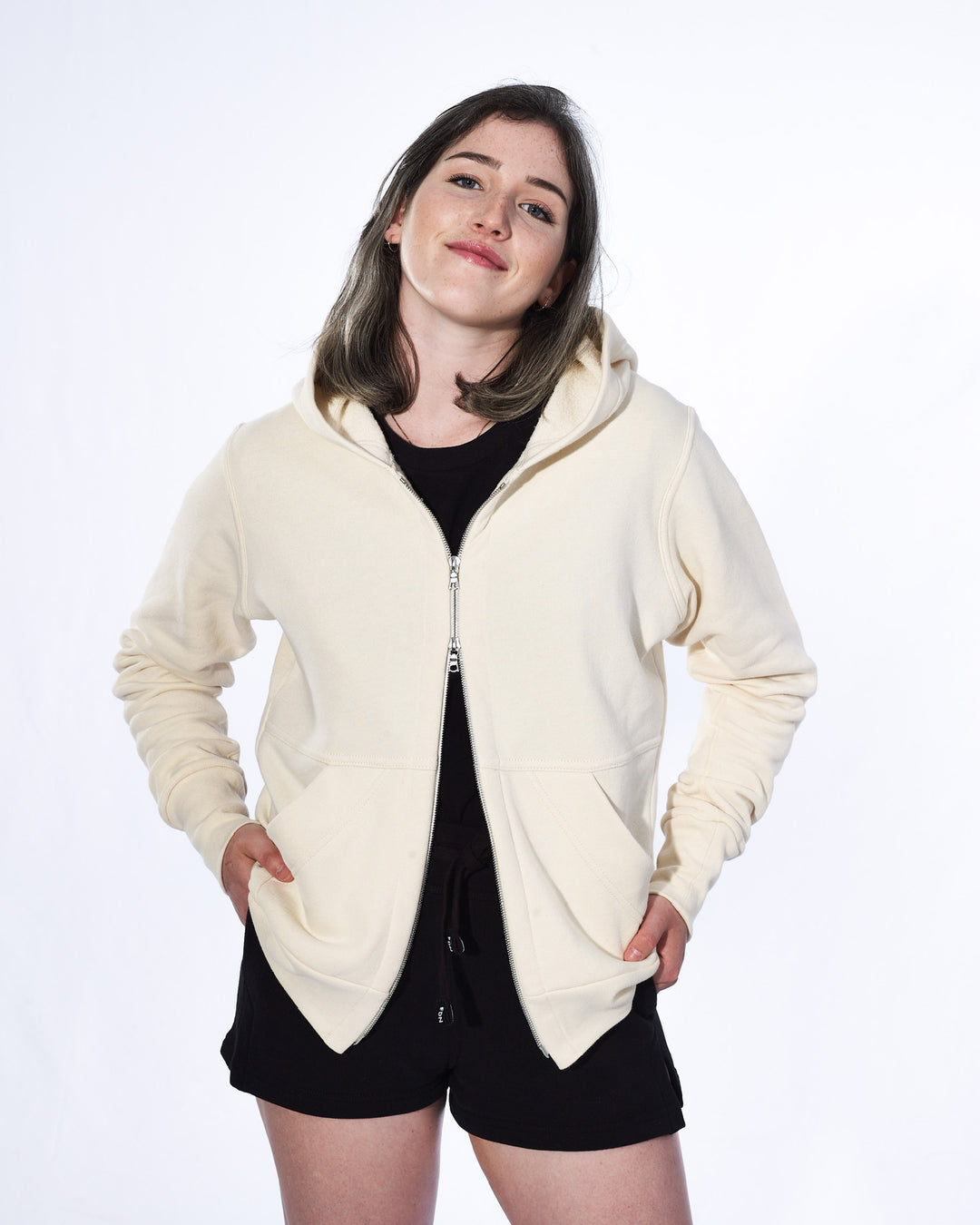 A girl with long sable hair standing in front of a white background. She is wearing a white cream zip-up hoodie, black t-shirt and black shirts. The hoodie has a two way silver zipper and two front pockets.