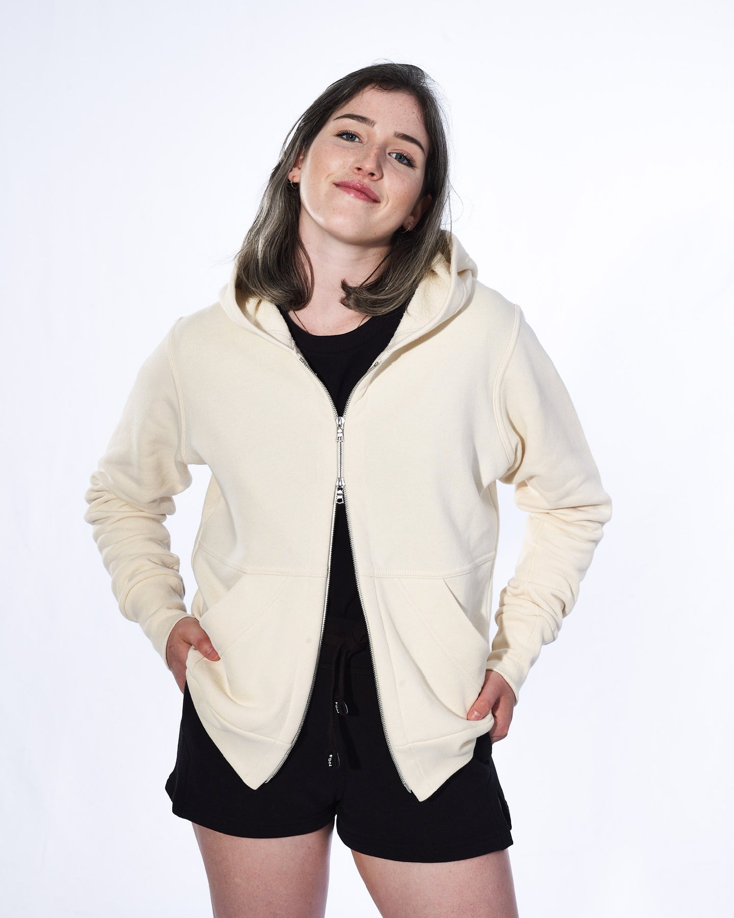 A girl with long sable hair standing in front of a white background. She is wearing a white cream zip-up hoodie, black t-shirt and black shirts. The hoodie has a two way silver zipper and two front pockets.