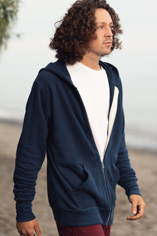 A man with curly long hair walking on the beach in overcast weather. Wearing a navy zip-up hoodie with a two way silver zipper and two front pockets.