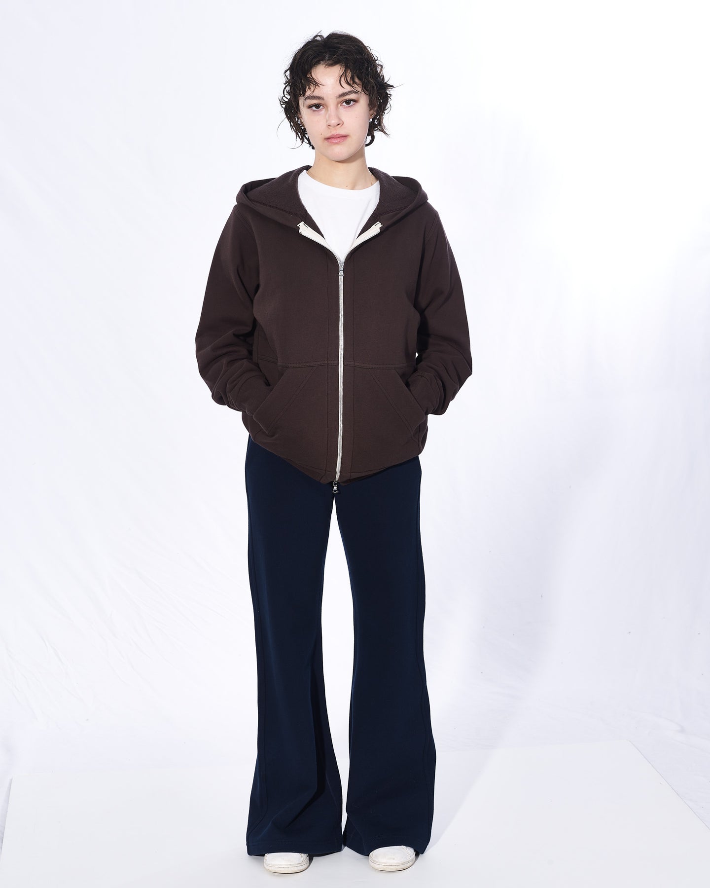 A girl with short brown hair standing with her hands in her hoodie pockets in front of a white background. She is wearing a dark brown zip-up hoodie, white t-shirt, navy bell bottom pants and white shoes. The hoodie has a two way silver zipper and two front pockets.