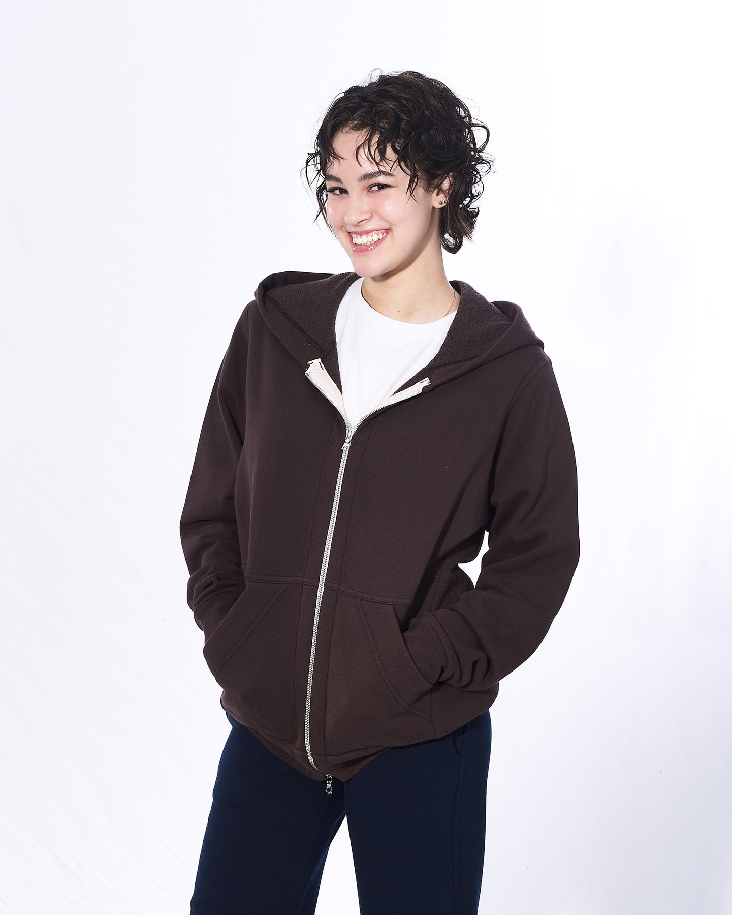 A smiling girl with short brown hair has her hands in her hoodie pockets in front of a white background. She is wearing a dark brown zip-up hoodie, white t-shirt and navy pants. The hoodie has a two way silver zipper and two front pockets.
