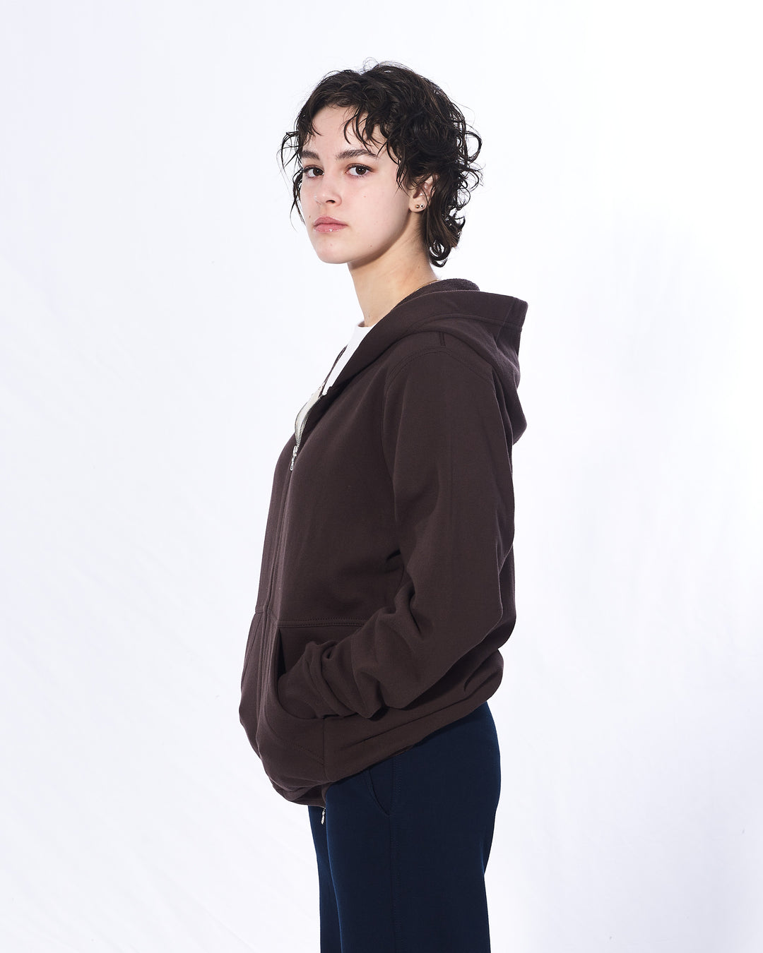 A girl with short brown hair standing sideways with her hands in her hoodie pockets in front of a white background. She is wearing a dark brown zip-up hoodie, white t-shirt and navy pants. The hoodie has a two way silver zipper and two front pockets.